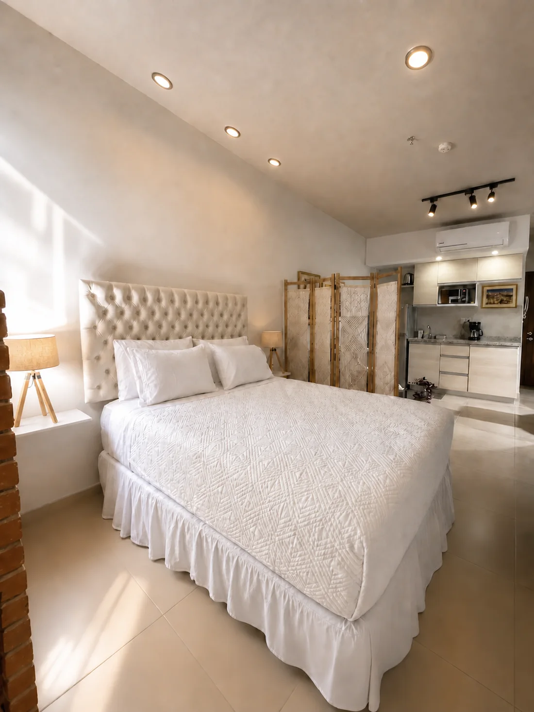Cama queen con cabecera capitonada y kitchenette al fondo